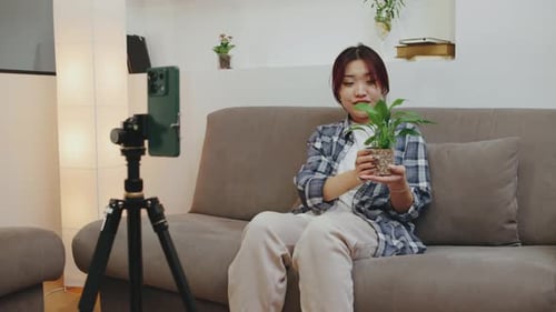Woman Smiling While Holding a Potted Plant During an Indoor Video Call in a Cozy Living Room at Home