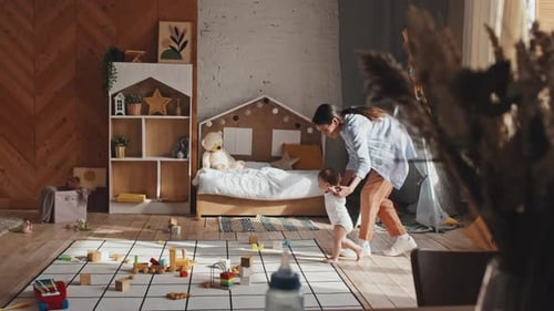 Baby Making First Steps On Floor, Supported By Mom, Toys Lying Around