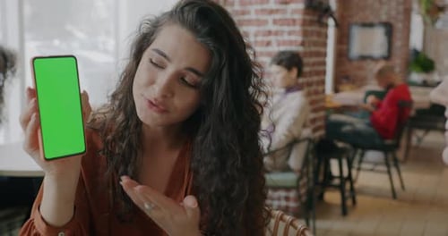 Beautiful Lady Pointing at Smartphone with Chroma Key Green Screen and Talking to Friend Indoors in