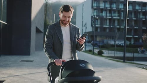 Smiling Young Handsome Father with Little New Born Baby in a Stroller Walking Use Phone at Sunlight