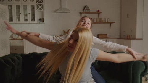 Fröhliche, familienliebende junge Mutter spielt Huckepack, süßes kleines lustiges Kind, Tochter, sitzt auf der Couch