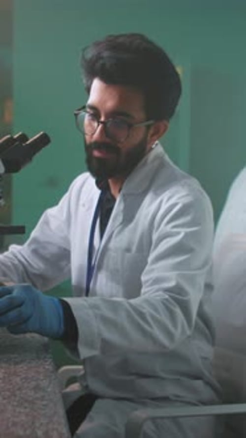 Ein lächelnder Mann, Wissenschaftler mit Brille und Schutzhandschuhen, der unter das Mikroskop schaut, hält eine Röhre