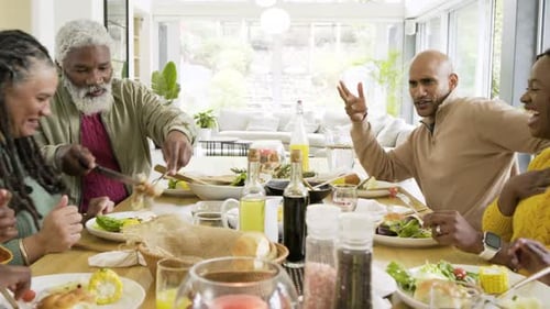 Diverse family gathering at dining table for meal, causing man scooping salad and passing bread