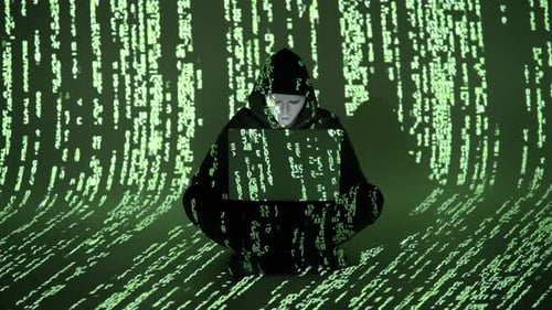 Cybercriminal in Mask and Hoodie Sitting with Laptop Amid Falling Green Code in Dark Digital Room