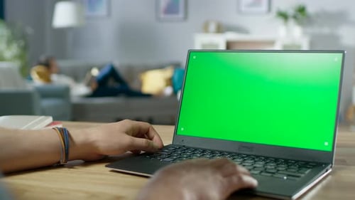 Over the Shoulder Shot of Mans Hands Typing on a Laptop with Green Mock-up Screen. In the Backgroun