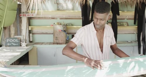African american man preparing surfboard on the counter of surf rental beach shack, slow motion