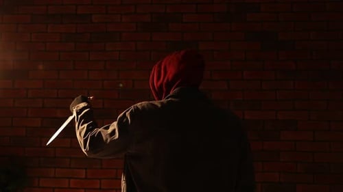 Criminal man in hood with knife near brick building outdoors