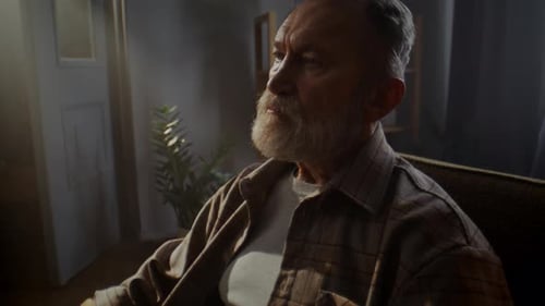 Upset Elderly Man with Grey Beard Sits in Armchair Feeling Lonely