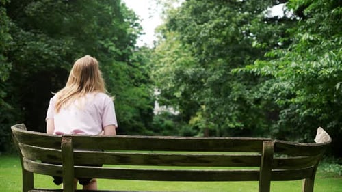 Nice and Slim Young Woman in Black Shorts Holding a Coffee Sits on a Park Bench Savoring Her Drink