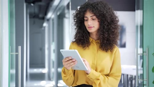 Young adult businesswoman is using digital tablet while standing in modern business office. Curly