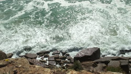 Like white foam, ocean water crashes across rocky coastline. Slow-mo
