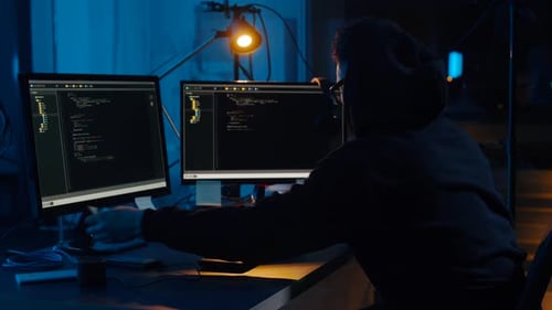 Man in hoodie types code on dual monitors