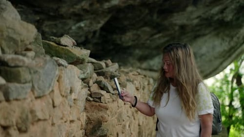 Mulher asiática sênior ativa com mochila explora os arredores da Gruta Rochosa em um smartphone envolvida em