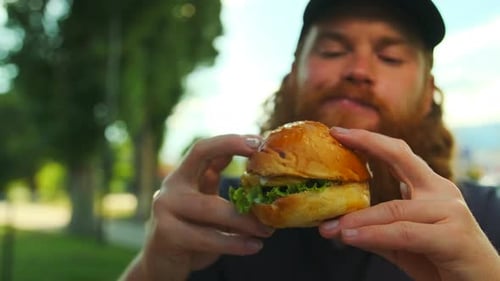 Young bearded man eating hamburger sitting in cafe outdoors. Junk street food. Close up