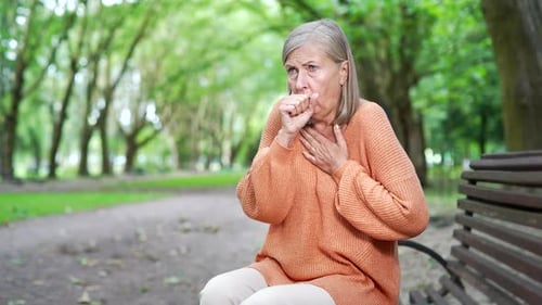 A sick senior female with gray hair coughing sitting on a bench on the street in an urban city park.