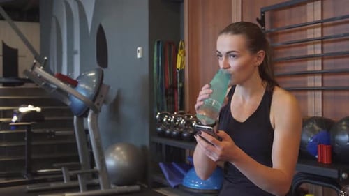 Woman Using Phone and Drinking Water in Gym