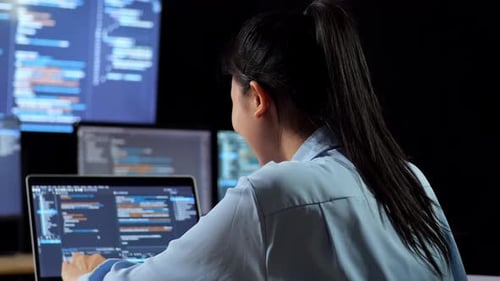 Vista trasera de una programadora asiática escribiendo código con una computadora portátil usando varios monitores