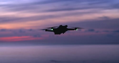 Silhouette of Modern Drone Hovering in the Air and Turning on Sea Background at Sunset Copter with