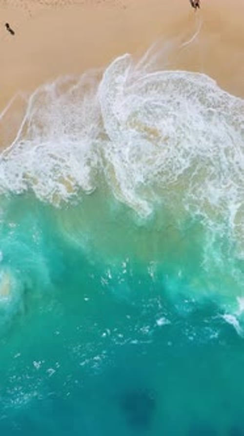 Vertical drone footage of turquoise ocean waves crashing on a sandy beach.