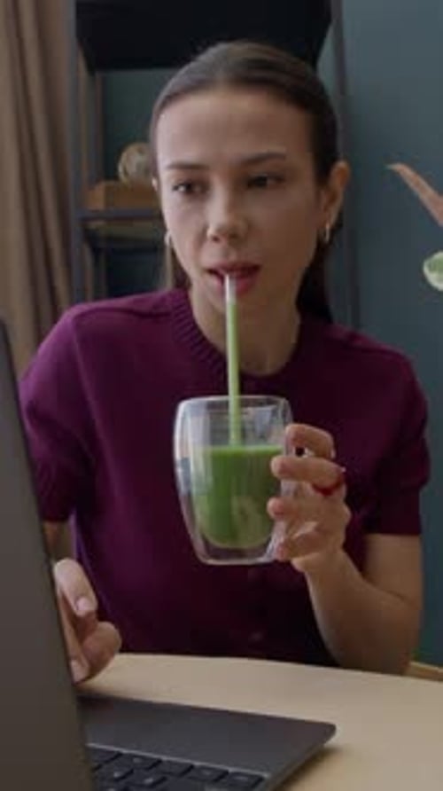 Young Woman Sips Smoothie While Working on Laptop