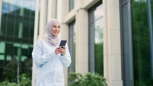 Young smiling muslim female employee in hijab is using a smartphone while walking on street near