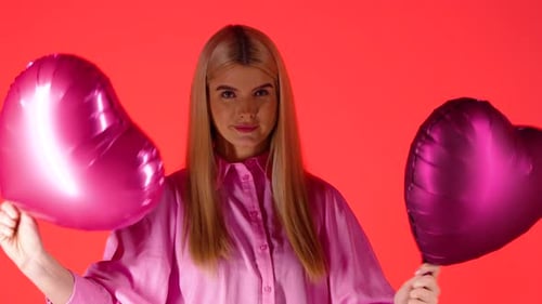 Pretty Blonde Woman Holding Waving Purple Heart Shaped Balloons Against Red Background, Studio Shot