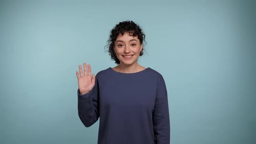 Lächelnde lockige Frau begrüßt winkende Hand und zeigt Hello Gesture blickt in die Kamera