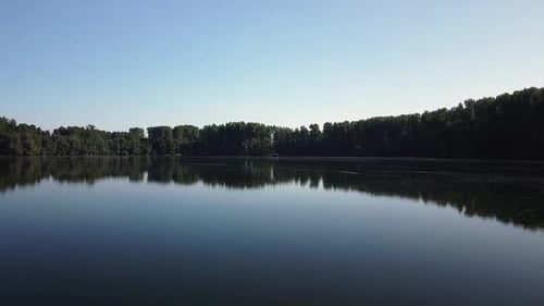 Drone flying close over the water of a dark eerie lake with reflections of trees and blue skies in E