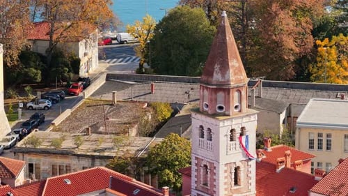 Aerial drone view of a tall church tower displaying the Croatian flag, surrounded by autumn colored