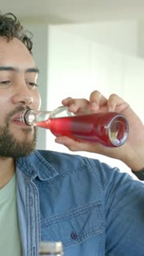 Vertical video: Bringing bottle to lips denim-clad man sipping red drink testing taste in kitchen