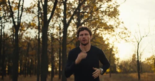 A Confident Man with Curly Hair and a Beard in a Black Sports Uniform Runs Along a Forest Path in an