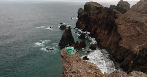 Aerial view of a man sitting at the edge of a dramatic cliff overlooking the ocean in Madeira, Portu