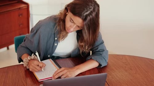 Young Woman Writing in Notebook with Laptop at Table