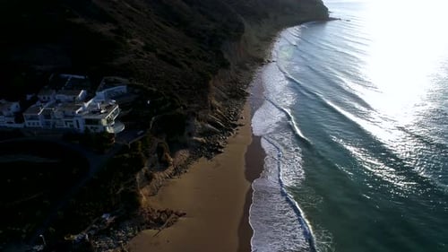 Aerial shot of waves crashing at beautiful beach with a big villa standing above the cliff