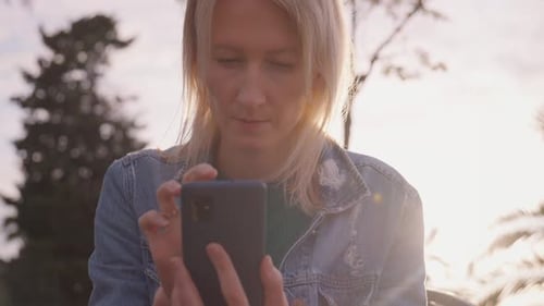 Woman Using Cell Phone in City Park at Sunset