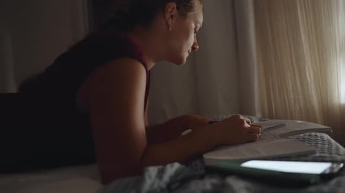At night, the young woman lies on the bed and works, copying a notebook.