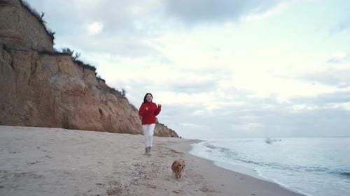 Beautiful Brunette Ukrainian Woman in a Red Sweater Runs Along the Sea with Two Small Dogs in Autumn