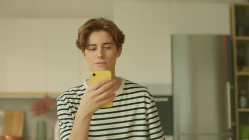 Moving Shot of Young Cute Caucasian Man Sitting at Table and Using Smartphone in Modern Kitchen Guy