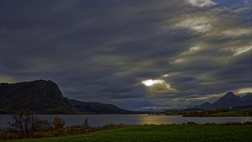 Panoramic view of a lake surrounded by mountains on a cloudy day in late autumn; penetrating sunbeam