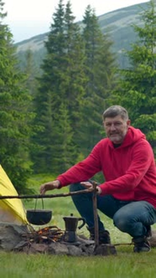 Man Cooking Over Campfire Near Tent in Mountain Meadow