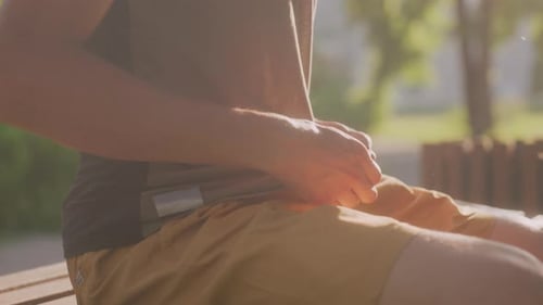 Man Adjusts Belt While Sitting Outdoors on Bench