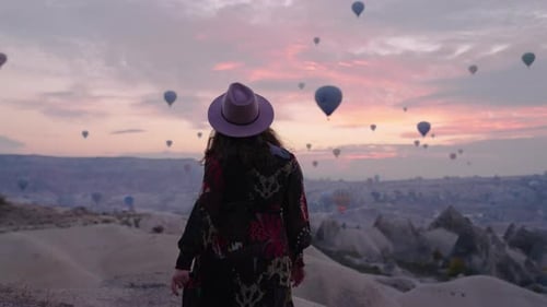 Girl In A Dress Watching Hot Air Balloons In The Sky Of Cappadocia In Turkey - wide shot