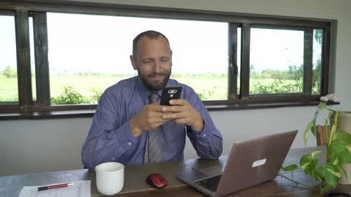 Young businessman texting on smartphone at table in his home with a cheerful smile
