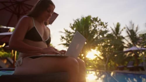 Junge Frau arbeitet aus der Ferne an einem Laptop, während sie sich beim Golden neben einem tropischen Swimmingpool entspannt