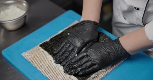 Un chef portant des gants en nitrile noir prépare des sushis dans la cuisine en étalant du riz cuit sur du nori
