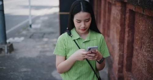 Asian woman looking at a map on mobile app while walking along a sidewalk next to a brick wall.