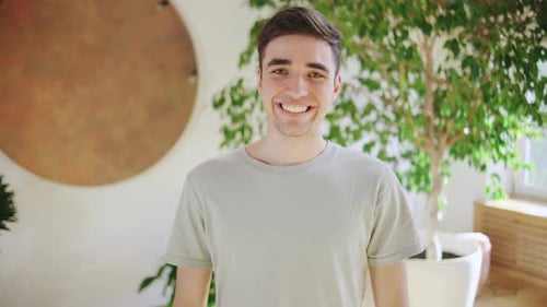 In a Bright Modern Room a Cheerful Young Man Stands Confidently Wearing a Casual Gray Shirt Exuding