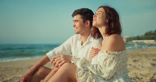 Happy Brunette Guy in a White Shirt Sits with His Brunette Girlfriend in a White Dress on the Beach