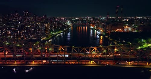 Multiple cars crossing the bridge at night. City lights reflecting in the river water.