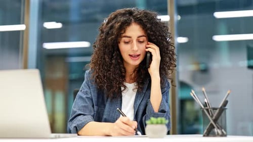 Young Businesswoman Talking on Phone and Taking Notes in Modern Office